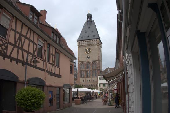 Blick durch eine Gasse auf einen hohen Turm mit schwarzem Dach. im unteren Drittel drei hohe Rundbogenfenster. links in der Gasse ein Fachwerkhaus. Den Weg zum hohen turm im Hintergrund säumen Sonnenschirem und Sitzgelegenheiten mehrerer Cafes.