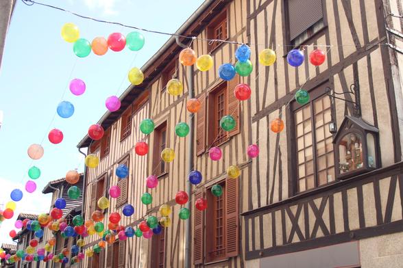 eine Häuserreihe mit französischem Fachwerk, die dunklen Holzbalken stechen aus dem hellocker der verputzten Zischenräume stark hervor.zwischen der Häuserreihe sind bunte Luftballons aufgehängt und gegen der Szenerie einen fröhlichen Charakter.