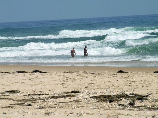Breiter Sandstrand, auf dem dnkler Tang liegt, mit Blick aufs Wasser, das in ca ein meter hohen Wellen an den Strand brandet. Zwei Bedegäste gehen gerade ns dunkekltürkisfarbende Wasser.