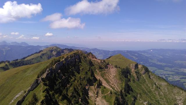 Bild von eiem Gipfel in die gegenäberligenden Bergspitzen. Es sind keline Pfade zum Anderen Gipfel sichtbar welche grüne Berwiesen zerschneiden und um Felsformationen herumführen. Der Himmel ist blau mit vereinzelten Haufenwolken, ganz im Hintergrund steht eine Dunstwand.