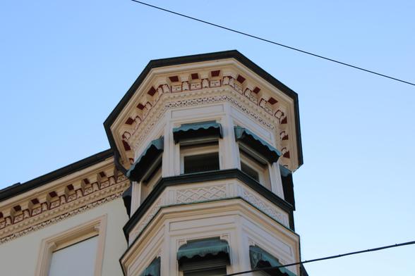 Bild ist nach oben in den blauen Himme geschossen und zeigt den das oberste Stockwerk eines Hauses mit einem sechseckigen Erker, der im neoklassizistischem Stil gestaltet ist. Der Erker zeigt uns vier Fenster, rechteckig in wagerechter Wweidrittelteilung und einer grünenen Haube oben, unter der sich der Rolladen verbirgt. Das Gebäude ist weiß gestrichen.