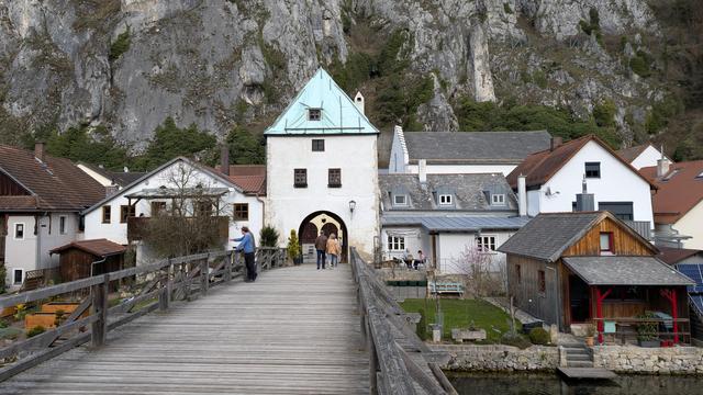 Turm mit Tordurchfahrt nach einer Holzbrücke, die wir im Vordergrund sehen. Der Torbogen ist rund hat die Höhe des unteren Viertels des Turmes, darüber drei Fenster, zwei im ersten Stock, eiens im zweiten Stock und dann ein spitz zulaufendes Dach. Es ist mit Plane abgedeckt, weil es reoariert wird. Rechts und links des Tores Wohnbebauung, dahinter eine Kalkfelswand.