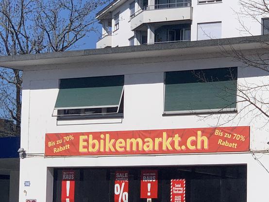 Blick auf eine Hausfassade, an dem ein Transparenz mit der Aufschrift ebikemarkt.ch, bis zu 70% Rabatt hängt. Als Alemanne lese ich das gemeinte E-Bike automatisch als Ebike, was dialektal dem Ortsnamen Ebikon entspricht.