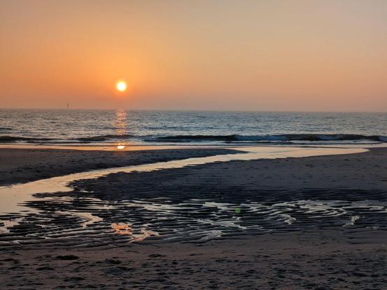 eine tiefstehende orange Sonne spiegelt sich im flachen Wasser am Sandstrand