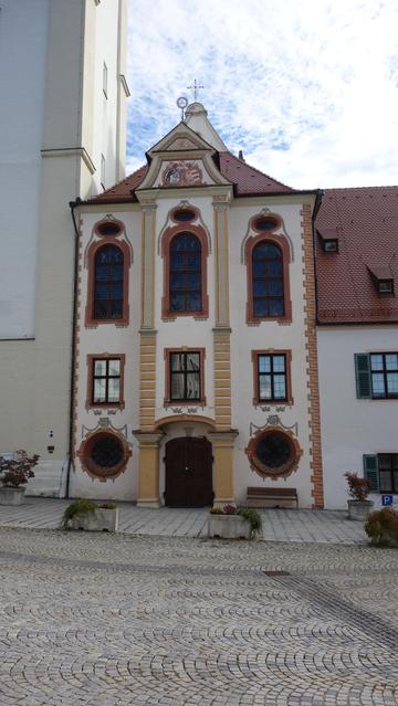 Stirnseite einer Kirche mit zwei Reihen a drei Fenstern nebeneinander und mit zwei angedeuteten Säulenkaptitellen voneinander getrennt. Die oberen länglichen Fenster sind mit braunem Rahmen eingefasst. unten links und rechts neben der Tür in der Mitte zwei runde Fenster. Links neben der Stirnseite schließt der Kirchtum an.