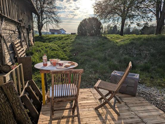 Auf einer kleinen Holzterrasse steht ein runder Tisch mit bunten Beinen und Essen draufn2 Stühle nAbendsonne scheint drauf 