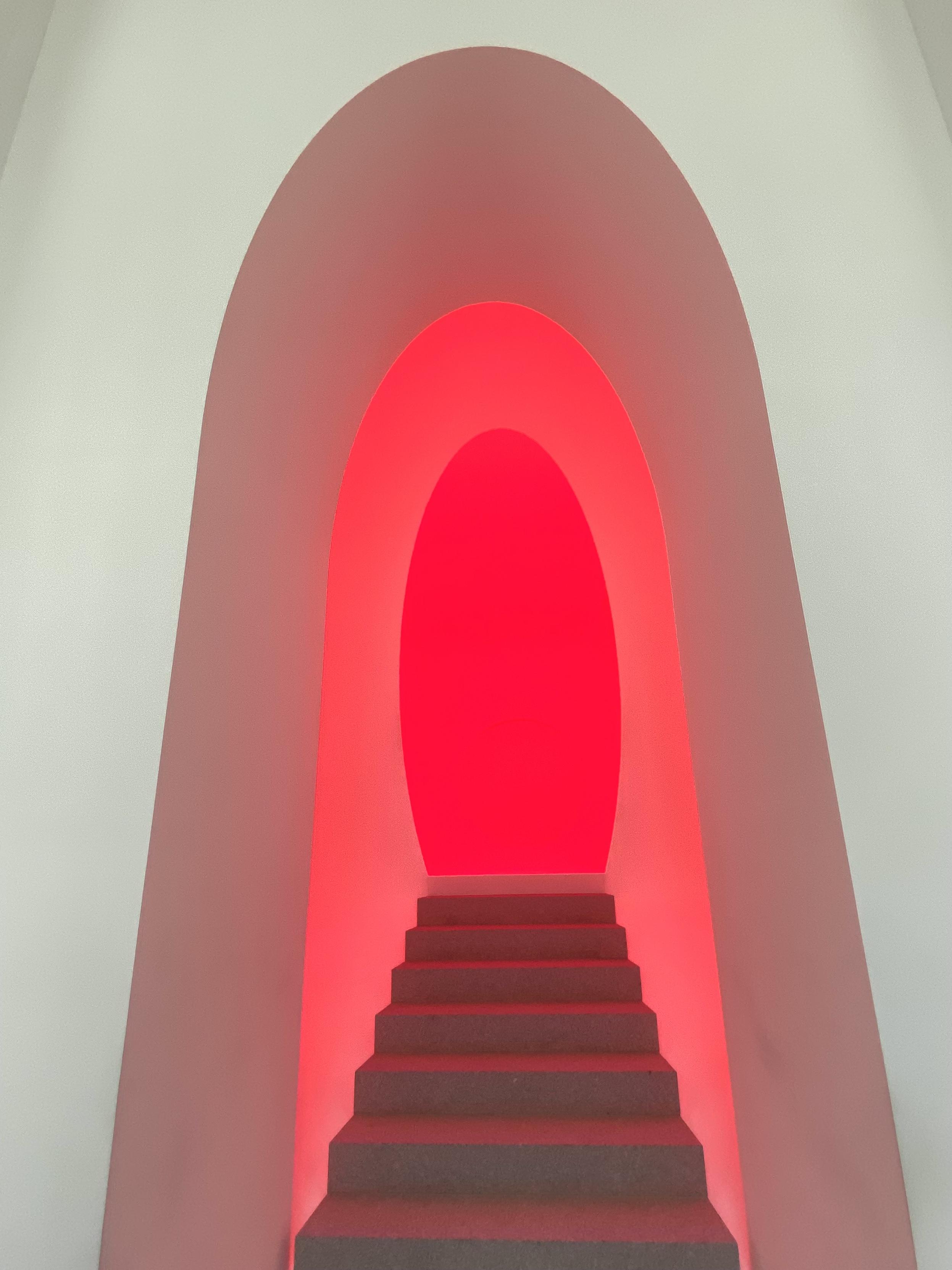 Eine schmale Treppe führt durch einen parabelartig gerundeten Eingang nach oben in rötliches Licht.