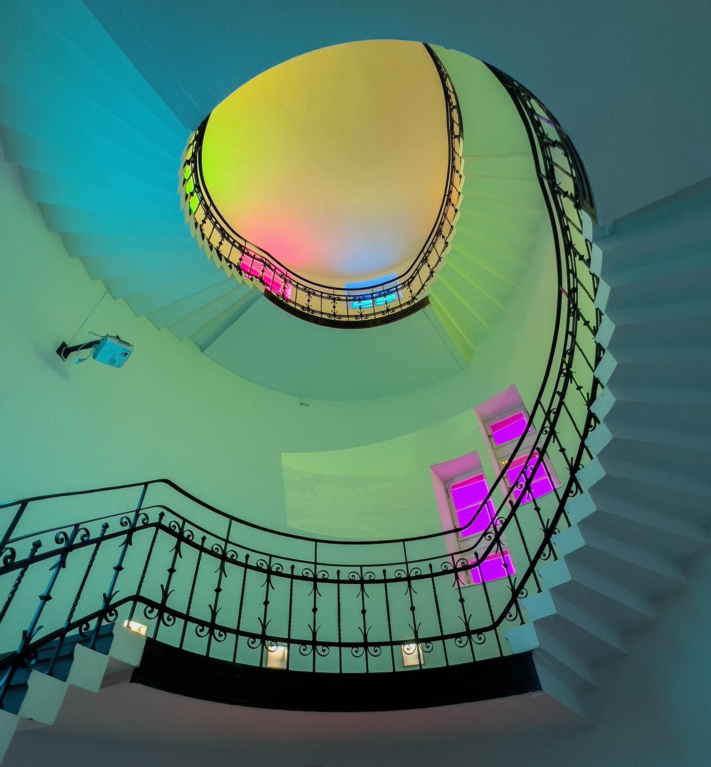 Eine beeindruckende Untersicht direkt in das Zentrum einer Wendeltreppe. Die hellen Stufen und das filigrane, dunkle schmiedeeiserne Geländer mit Schnörkeln bilden eine perfekte Spirale, die nach oben führt. Die Wände sind durch eine Lichtinstallation in ein sanftes Farbspiel aus Mintgrün und Türkis getaucht, das zur Decke hin in ein warmes Gelb übergeht. An der rechten Wand leuchten vier übereinander angeordnete Fenster in einem intensiven, leuchtenden Magenta.