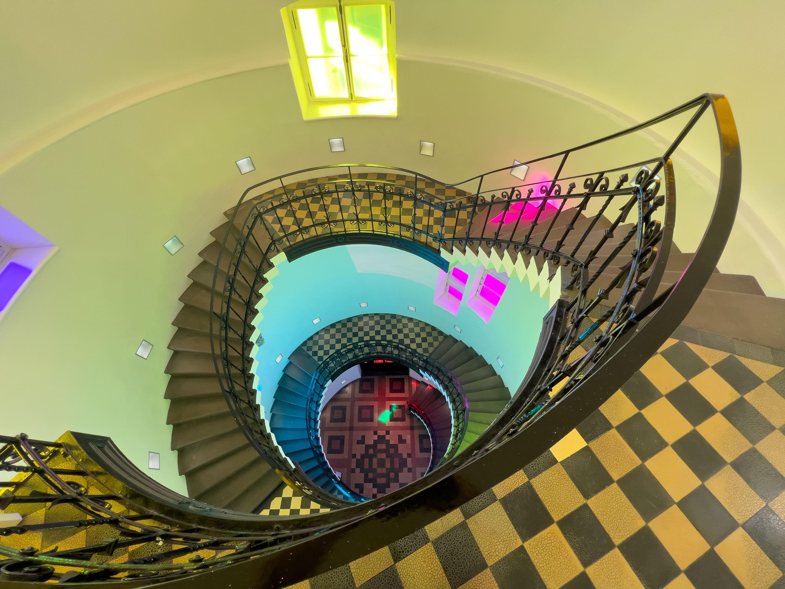 Eine dramatische Aufsicht von oben in den Treppenlauf einer Wendeltreppe. Die dunklen Stufen und das verzierte Metallgeländer winden sich kreisförmig in die Tiefe. Die Szene ist von bunten Lichtprojektionen geprägt: Auf den Böden und Wänden sind Muster in Magenta, Grün und Blau zu sehen, darunter ein auffälliges schwarz-weißes Schachbrettmuster auf einem Treppenabsatz. Am tiefsten Punkt der Spirale leuchtet eine Tür in kräftigem Rot. Die Wände sind in kühles Mintgrün getaucht.