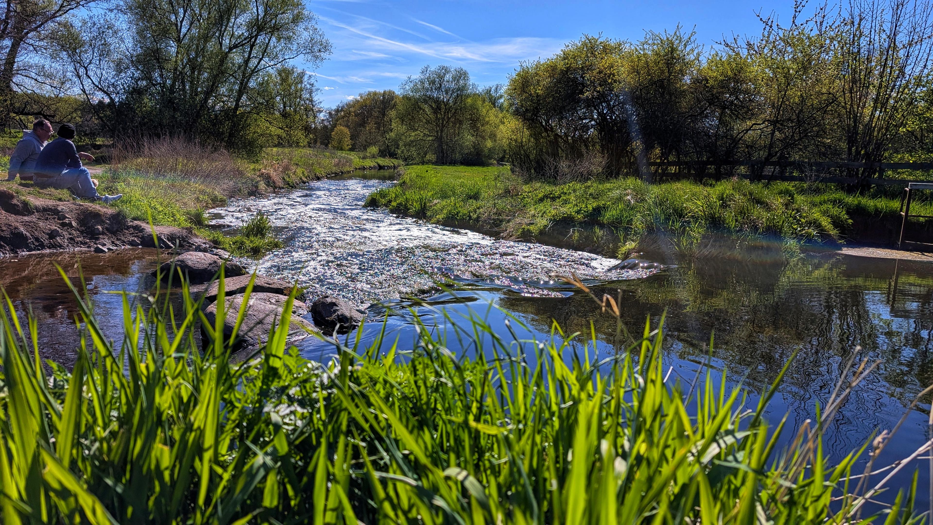 Eine Landschaft mit einem fließenden Fluss, grüner Vegetation und Bäumen unter einem blauen Himmel. Im Vordergrund sind unscharf grüne Grashalme zu sehen. Ein Fluss fließt durch die Bildmitte, wobei auf der linken Seite zwei Personen auf einem felsigen Ufer sitzen. Die Ufer sind mit dichtem grü grünem Gebüsch und hohen Bäumen bewachsen. Der Himmel ist hellblau mit einigen dünnen, weißen Wolkenstreifen.