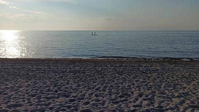 Zwei Standup-Paddler als Silhoutte ca. 100 Meter entfernt vom Ufer auf spiegelglattem Meer im Abendlicht. Das Ufer ist ein ca. 30 Meter breiter Sandstrand, dei Spuren im Sand werden duch dsAbendlich von der Linken Seitehervorgehogebn. Himme und Wasser blau, links reflektiert sich noch die Sonne mit einem Glitzern auf der Wasseroberfläche. Der Himmel trägt weiße Schleierwolken.