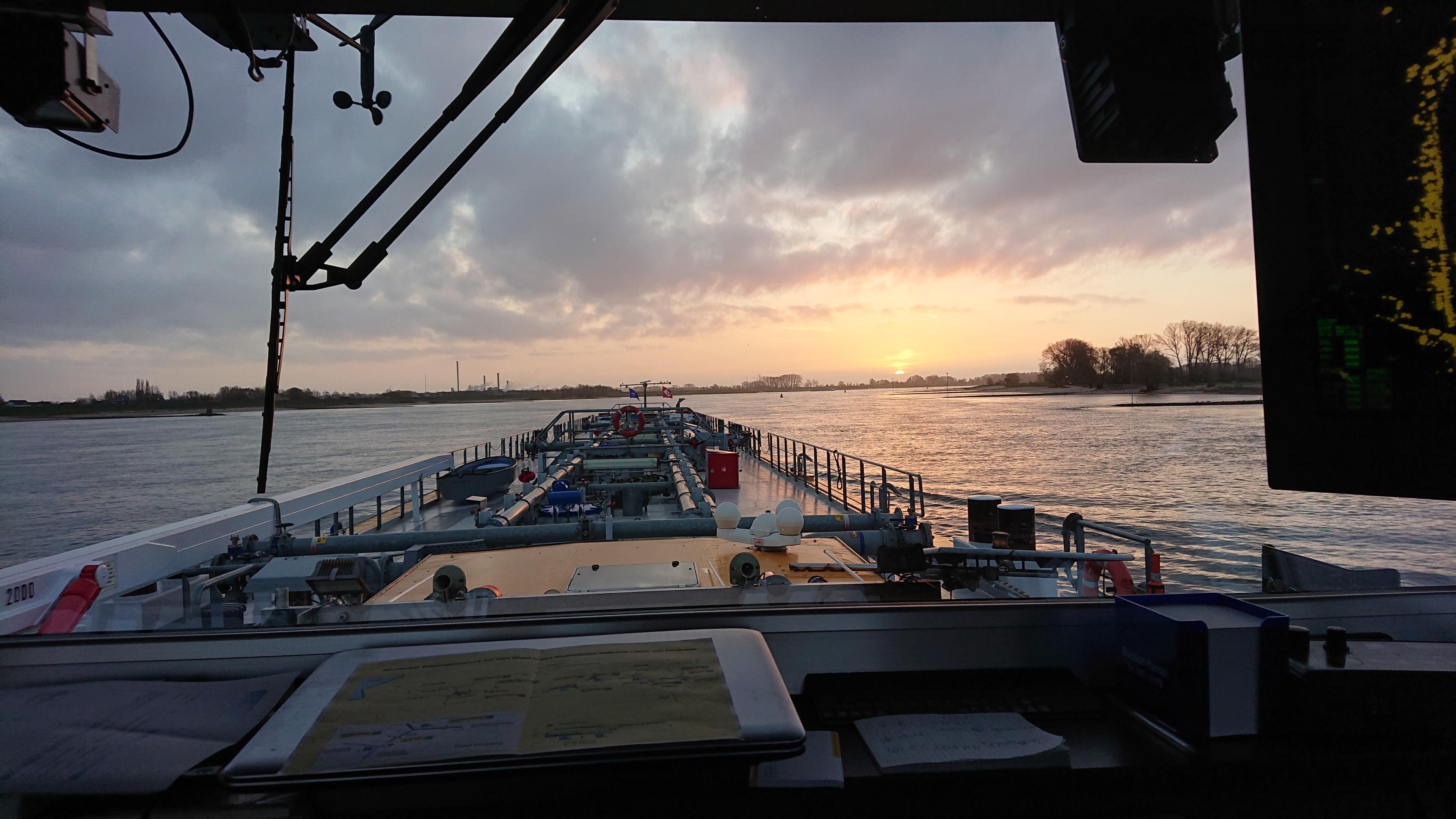 Aussicht aus dem Steuerhaus eines Tankmotorschiffes während der Fahrt auf einem Fluß, dem Sonnenuntergang oder -aufgang entgegen.