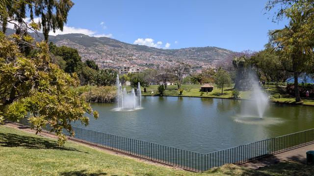 Zwei Springbrunnen in einem Park, links vorne hohe Bäume, im Hintergrund die bewohnten Hügel von Funchal in der Mittagssonne, darüber blauer Himmel und ein paar weisse Wolken über den  Gipfeln.
