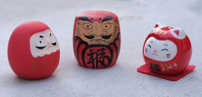 auf einer weißen Fläche stehen drei kleine Daruma Figurenn1x matt Vermillionrot und einfache Bemalungn1x fast zylindrisches Holz und aufwändig bemaltn1x glänzend rot mit Katzengesicht und kleinen spitzen Ohren