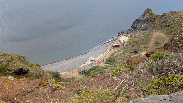 Bild zeigt eine Fotografie ca 100 Meter Höhenunterschied die Klippen hinunter auf einen kleinen Standabschnitt, der mit einem Gebäude, Parkflächen und Gastbereich ausgebaut ist. Auf dem Dach des Gebäudes steht "Welcome To Paradise", der Strand besteht aus groben Kieselsteinen. Der Wellengang ist minimal, das Wasser klar und leicht türkis. Es befinden sich sehr wenig Menschen unten am Strand. Die Klippen sind mit allerlei Grün wie Kakteen, Sukkulenten und anderem Kraut bedeckt. Rechts unten befi…