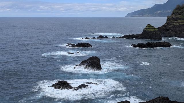Eine Kette bestehend aus vier Lavafelsen ragen schwaz aus dem Meer und werden von dem Wellen Schaumig weiß umspült. Im Hintergrund rechts dann die Insel mit einer steil aufragenden Steilküste, ebenfalls schwarzes gezacktes Lavagetein. Meer ca. einen Meter hohe Wellen, die sich an den Felsen brechen.