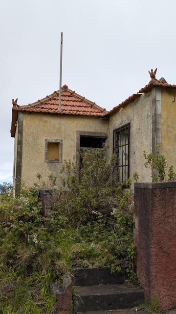 Einstöckiges kleines Haus mit Kaffeemühlenfibel, dessen vier nach unter laufende Ortgänge mit dein einem besonderen eine Figur tragenden Ortgangziegel abschließen. Vor der Tür und dem vergitterten Fenster wächst schon Gebüsch.