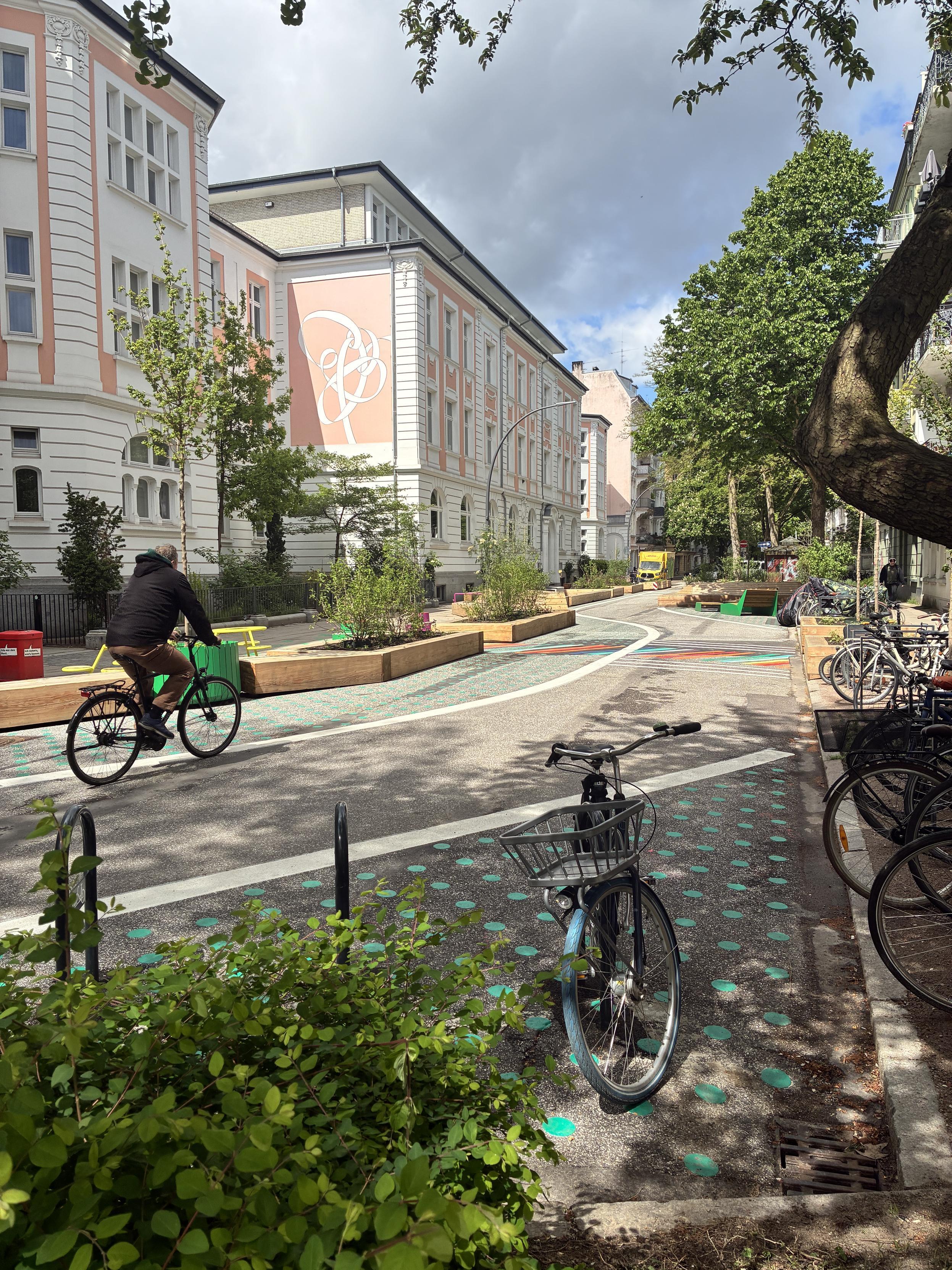 Foto der umgebauten Straße. Statt Autofahrbahn schlängelt sich ein breiter Radweg durch das Wohngebiet. In den neuen Beeten wachsen Pflanzen. Im Hintergrund hohe Bäume alten Bestands.
(Quelle: @hochbahn@norden.social)