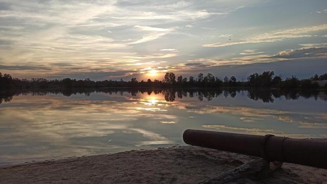 Die Sonne geht an einem See unter und färbt alle kleinen Wölkchen am Himmer rosa/orange. Diese spiegeln sich alle im stillen Wasser des Sees als Kopie des Himmels. Umrandet wird das durch die Spiegelgung der Bäume im See. Rechts im Vordergrund am sanigen Ufer führt ein Rohr in den See.