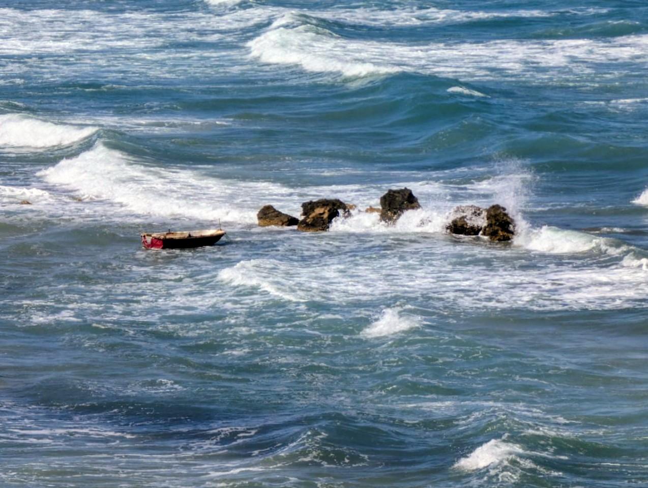 Du blickst auf ein aufgewühltes Meer, blaues Wasse, schäumende Gischt. Mittendrin ragen drei Felsen raus, und ein kleines rotes Ruderboot.