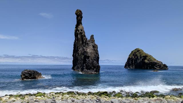 Drei Felsen ragen un Ufernähe aus dem Meer. Der Felsen in der Mitte ragt ca. 20 Meter mit einer Doppelspitze nach oben, wobei die rechte Spitze nur ein oder zwei Meter hoch ist. Eline Welle läuft auf den Kiesstrand auf und wirft die Gischt Recht im Bild nach oben. Die drei Felsen heben sich schwarz und kontrastreich vom blauen Himmel ab.