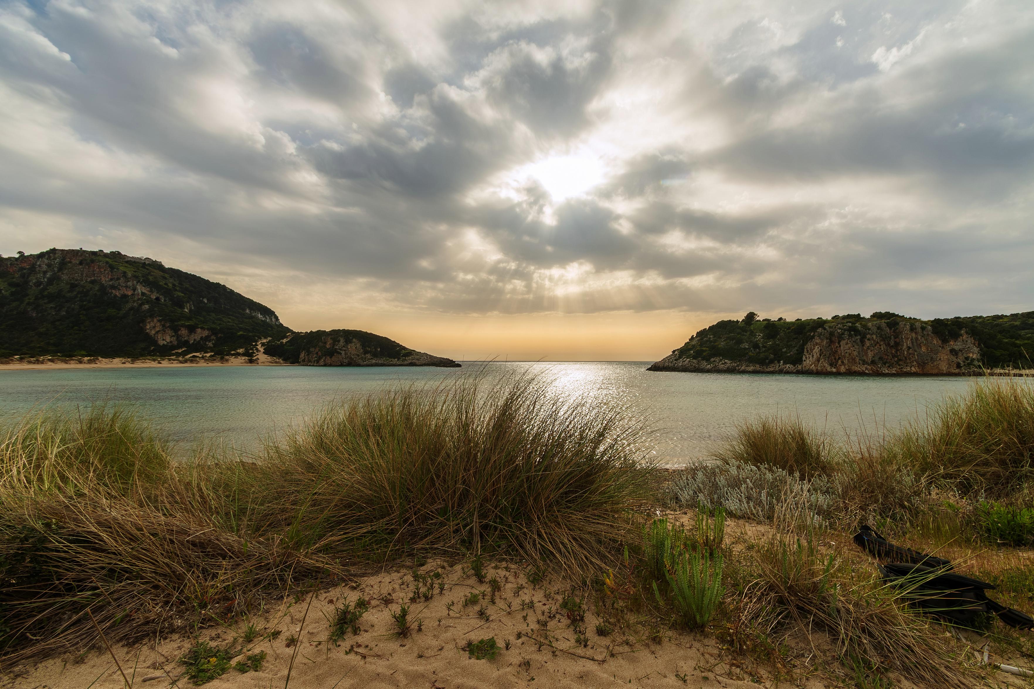 Die Aufnahme zeigt eine malerische Küstenlandschaft bei Sonnenuntergang oder Sonnenaufgang. Das Licht bricht durch die Wolken und wirft Strahlen auf das ruhige Meer. Im Vordergrund wachsen hohe Gräser und Büsche auf sandigem Boden, die eine natürliche Barriere zum Wasser bilden. Auf der linken und rechten Seite erheben sich bewaldete Hügel, die die Bucht umrahmen.