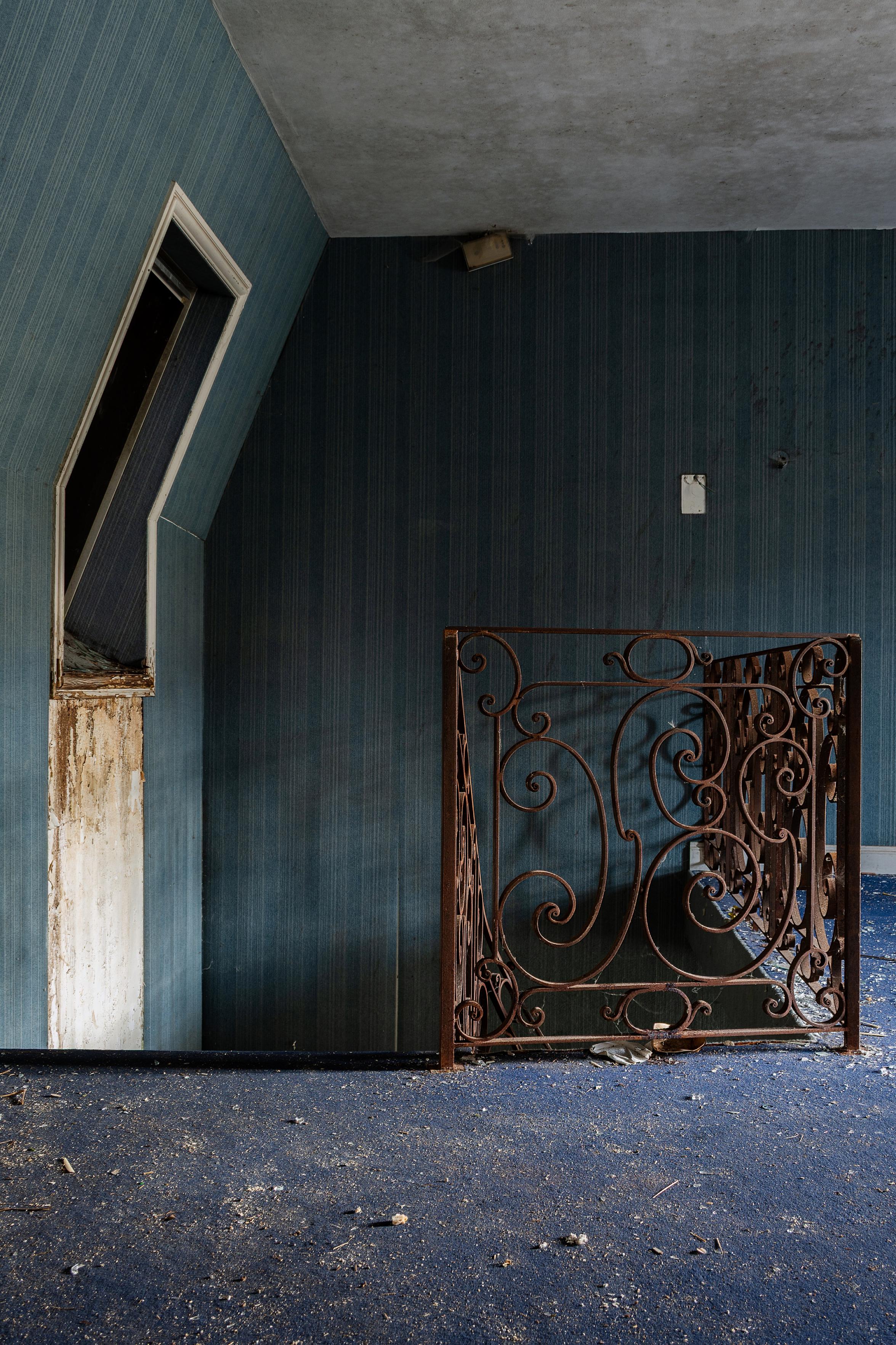 Das Bild zeigt ein verlassenes Zimmer mit einer schrägen Dachgaube und einem verzierten Metallgitter.

Im Bild ist ein Raum zu sehen, der auf Verfall und Vernachlässigung hindeutet. Die Wände sind mit einer blau-grauen, vertikal gestreiften Tapete versehen, die an einigen Stellen abblättert und beschädigt ist. Im Vordergrund liegt ein blauer Teppich, der mit Staub, Schmutz und kleinen Trümmerteilen übersät ist. Links im Bild befindet sich eine ungewöhnliche, schräg nach oben verlaufende Dachgaube mit einem schmalen Fensterrahmen, der von vergilbtem Holz umgeben ist. Der Fensterrahmen selbst zeigt ebenfalls deutliche Verfallsspuren. In der Mitte des Raumes steht ein kunstvoll geschmiedetes Geländer oder Gitter aus rostbraunem Metall mit filigranen Schnörkeln und Spiralen. Dieses Objekt steht auf dem Teppich und ragt in die Höhe. Im oberen Bereich des Bildes ist die Decke zu sehen, die ebenfalls einige Flecken und Unregelmäßigkeiten aufweist.