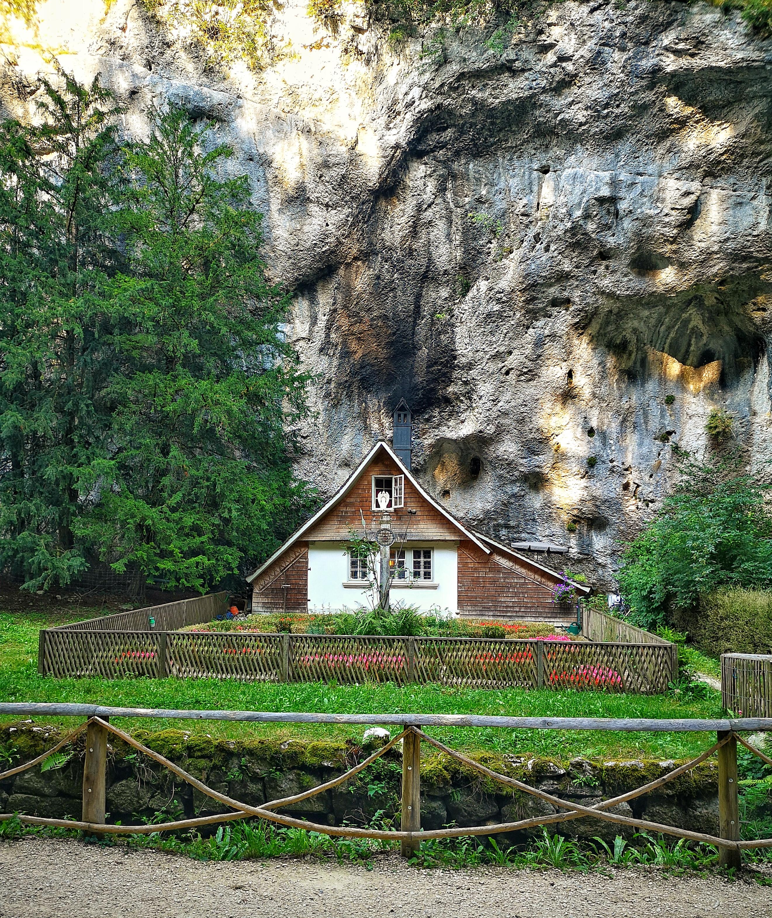 Ein winzig kleines Haus mit  kleinem umzäunten Garten inmitten von Wald vor einer überragenden Felswand.