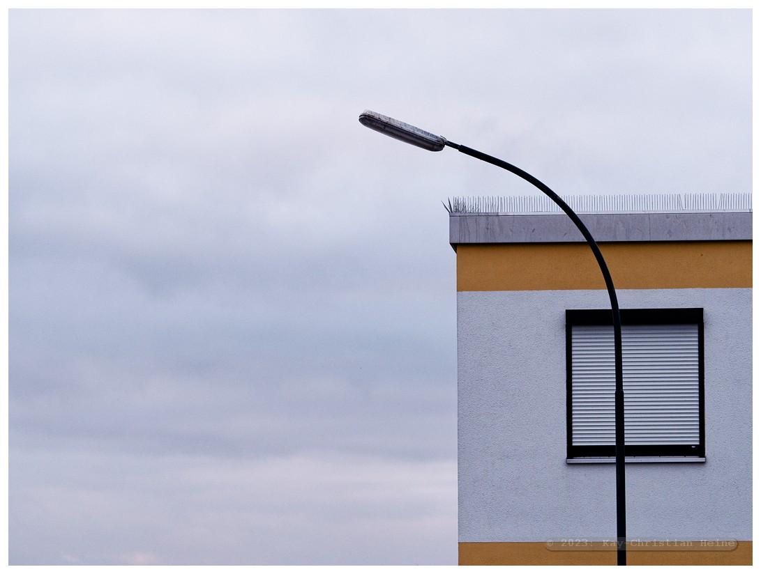 Rechte Bildhälfte: Ein Straßenlaternenmast mit gebogenem Arm ragt vor einem Gebäude mit Flachdach, weißer Fassade und gelben Streifen oben und unten in den Himmel. Das einzig sichtbare Fenster des Gebäudes ist mit einem heruntergelassenen Rollladen verschlossen. Auf der Dachkante sind Abwehrstacheln gegen Vögel angebracht. Der Himmel ist bewölkt. Die linke Bildhälfte ist bis auf den Himmel leer.