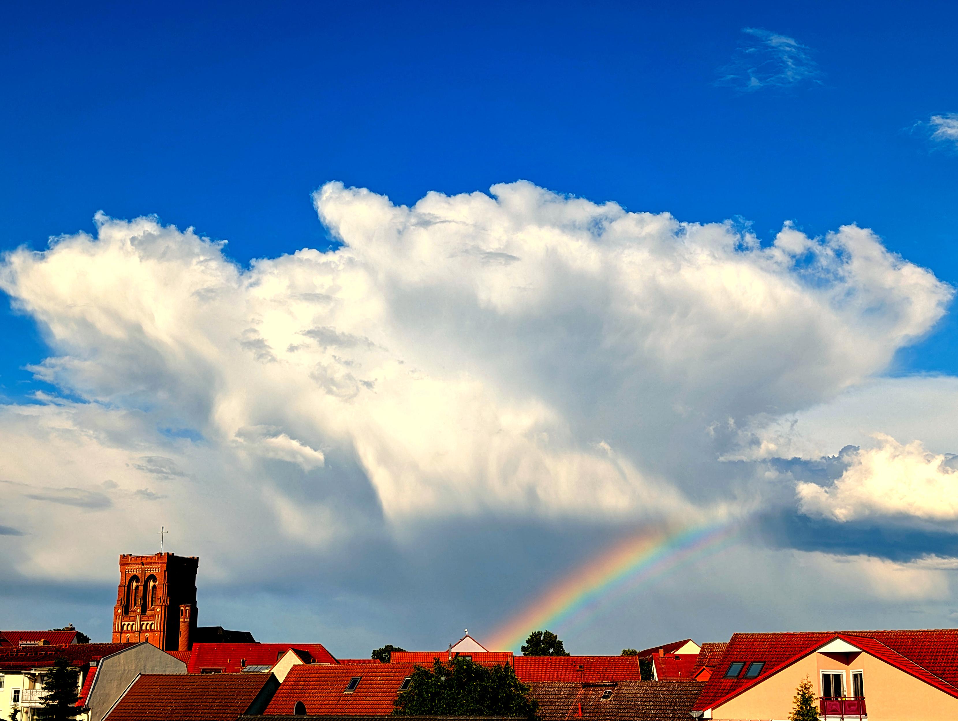 Das Bild zeigt eine Regenwolke mit leichter Amboßform vor blauem Himmel. In ihrem Schatten ist ein Regenbogen zu sehen. Im Vordergrund unter der Wolke die zumeist roten Dächer einer Stadt, aus denen ein Kirchturm mit zinnenbewehrtem Flachdach herausragt.