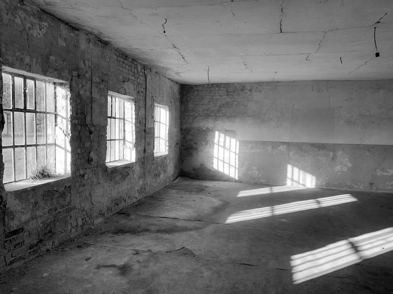Man sieht einen leeren Raum. Durch drei vergitterten Fenster leuchtet die Sonne hinein, so das Schattenbilder der vergitterten Fenster an der Wand und auf dem Boden zu sehen sind.