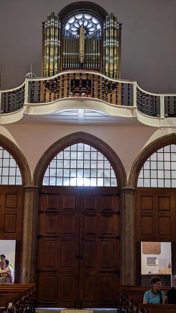 Blick auf eine Empore in einer Kirche, auf der eine Orgel steht. Hinter der Orgel ein rundes Fenster mit Rosette, welches von den zwei Seitentürmen eingerahmt wird. Unter der Empore drei von einem Gitter unterteilten Fenster, die gotisch spitz zulaufen.