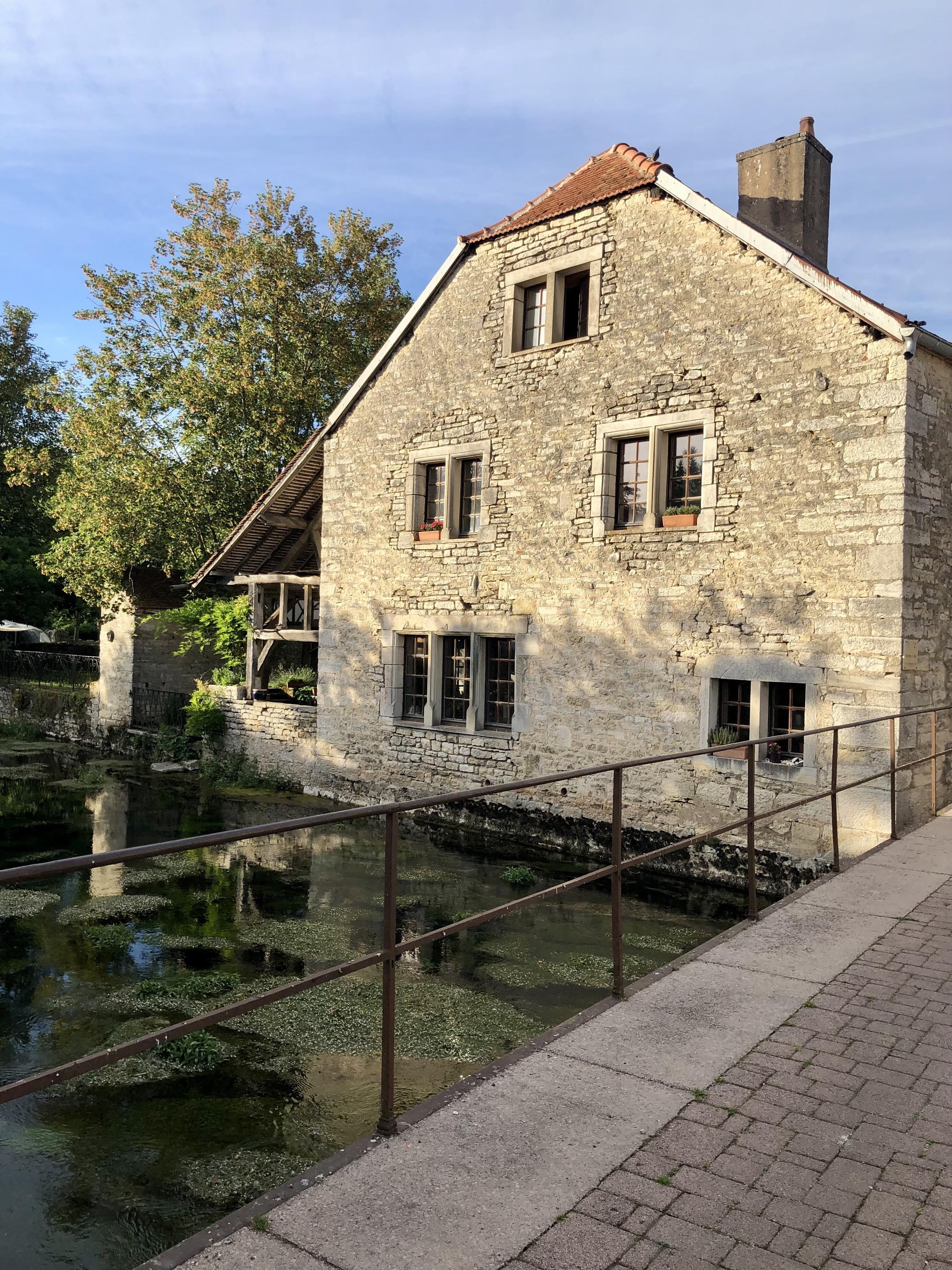Das älteste Haus des Ortes direkt am Fluss. Bruchstein, gelblich. Eine Terrasse am Wasser. Aufgenommen von einer Brücke.
