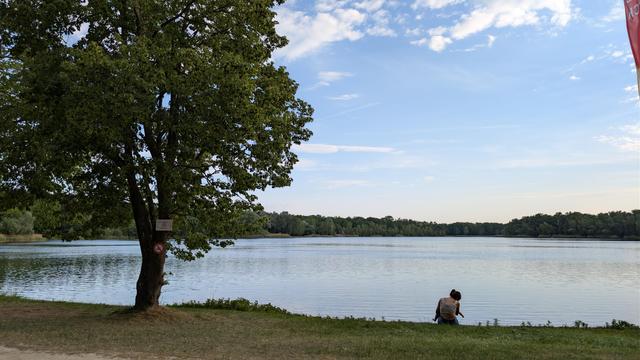 Blick auf einen See mit grüner Wiese alls Uferstreifen. Den See hinten begrenzen hohe grüne Laubbäume. Der Himmel ist blau, ein paar Wölkchen oben am Himmel. Links am Ufer ein Baum mit kugeliger dichtgrünbelaubter Krone. 5 Meter daneben sitzt eine Frau mit Rücken zum Fotografierenden und hat ein kleines Kind im Arm.