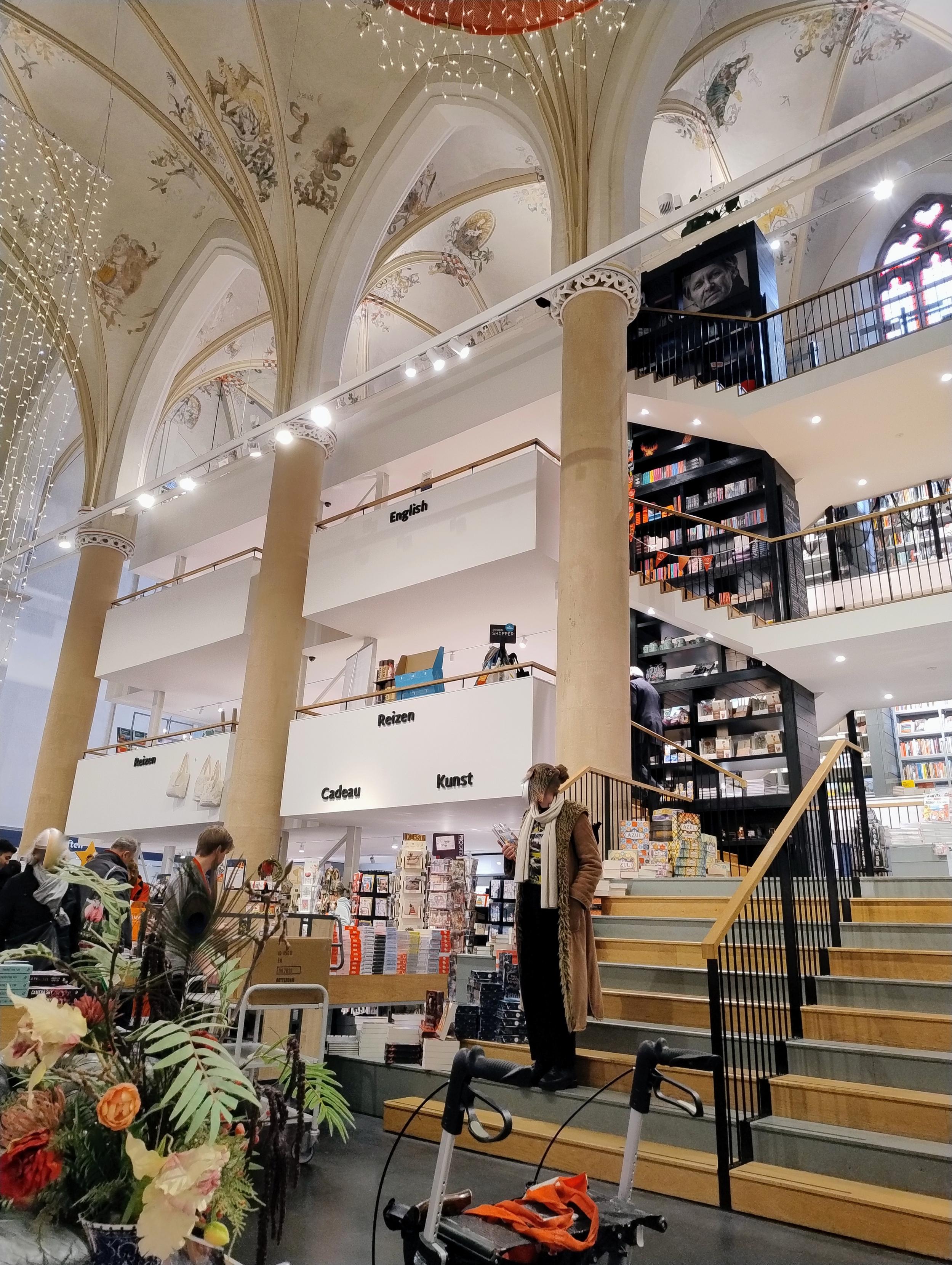 Blick oben auf das Gewölbe Richtung Seitenschiff erinnert großen Kirche. 

Treppen führen nach oben, es sind zwei Etagen in Seitenschiff eingezogen. Regale voller Bücher überall, im Erdgeschoss auch Ständer mit Karten und ähnlichem.

Leute laufen herum.

Das Gebäude ist innen weiß mit beugen Akzenten, an der Decke ist noch die alte Bemalung, im Hintergrund ist ein altes Kirchenfenster zu erkennen. Weihnachtliche Beleuchtung hängt.