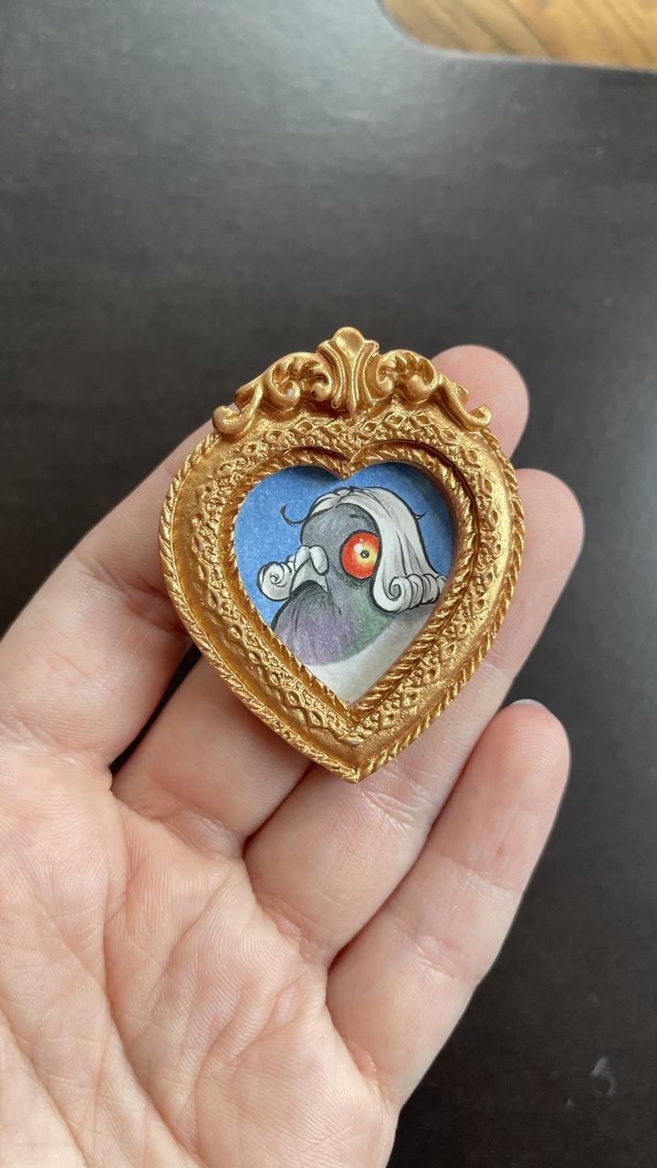 In einem herzförmigen goldenen Schnörkelrahmen, der auf meine Fingerspitzen passt, ist eine kleine Illustration einer Taube mit weißer locken Perücke. Er guckt blöde.