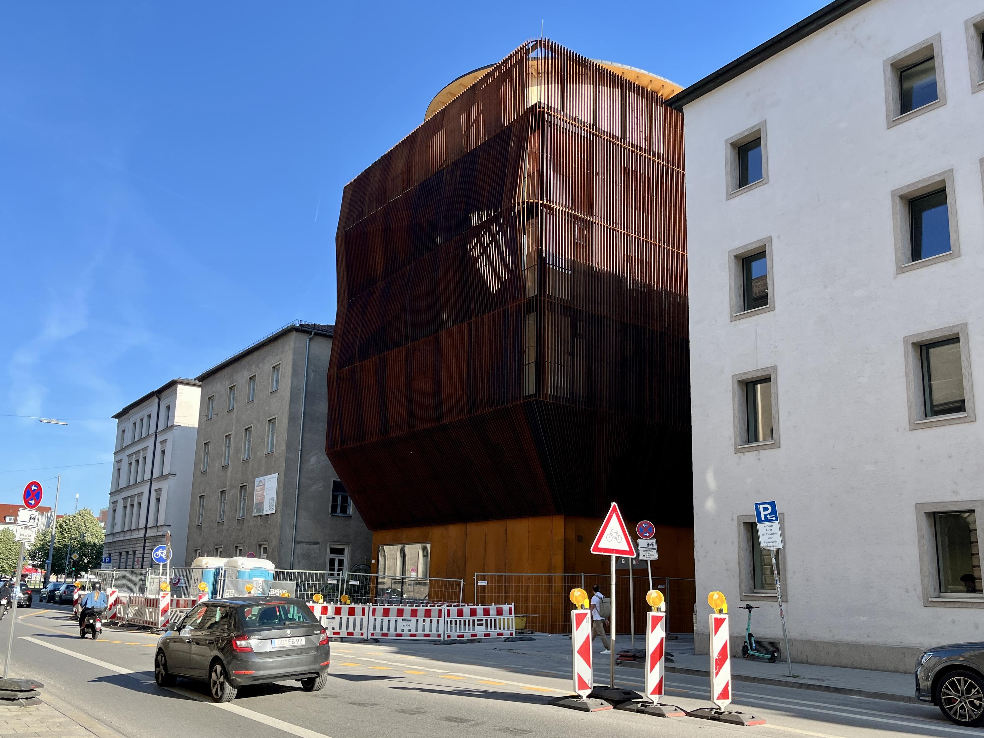 In einer durchschnittlich aussehenden Häuserreihe befindet sich ein auffälliges neues Gebäude mit einer unregelmäßig vorgewölbten Fassade aus rostroten Stabelementen.