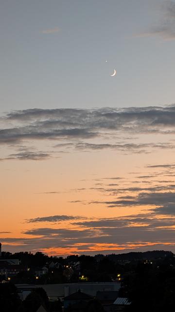 Der Mond und die Venus eng zusammen am orangen und mit locker mit vereinzelten Wolken bedeckten Abendhimmel, unten leuchten die Straßenlaternen einer Kleinstadt.
