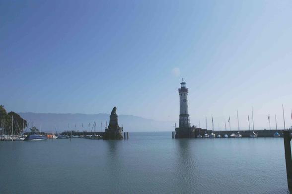 Blick von der Hafenmole auf die Hafeneinfahrt des Lindauer Bodenseehafens. Rechts der einzige Leuchtturm Bayerns, links der Einfahrt thront  eine Figur des bayerischen Löwens auf einem Sockel. Es ist morgens um ca. 9 Uhr an einem sonnigen Tag, die Berge im Hintergrund über Bregenz wie z.B. der Pfänder liegen im blauen Dunst. Das Wasser im Hafen reflektiert den blauen Himmel.