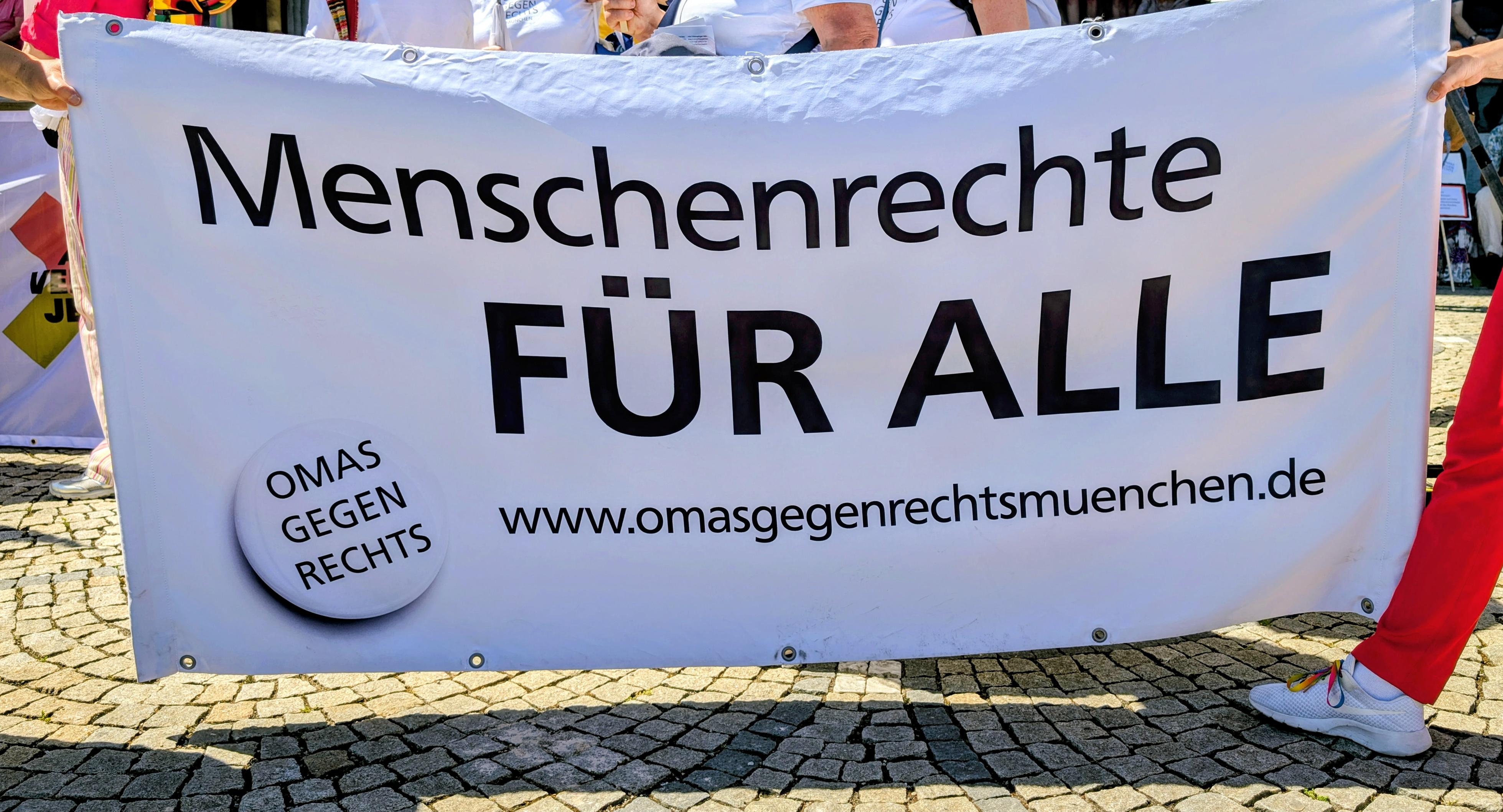Ein Transparent aus Stoff auf einer Demonstration. Mit schwarzer Schrift auf weißem Grund steht darauf: MENSCHENRECHTE FÜR ALLE. OMAS GEGEN RECHTS. www.omasgegenrechtsmuenchen.de
Im Hintergrund sieht man Arme und Beine der Personen, die das Transparent tragen.