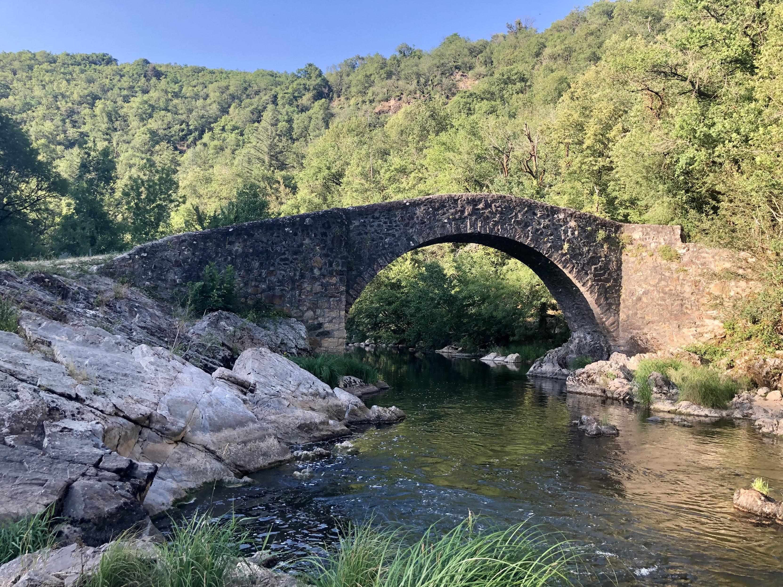 Eine steinerne Bogenbrücke in wilder Landschaft. Felsen und Bäume. Gleißendes Licht, ein Flusslauf.