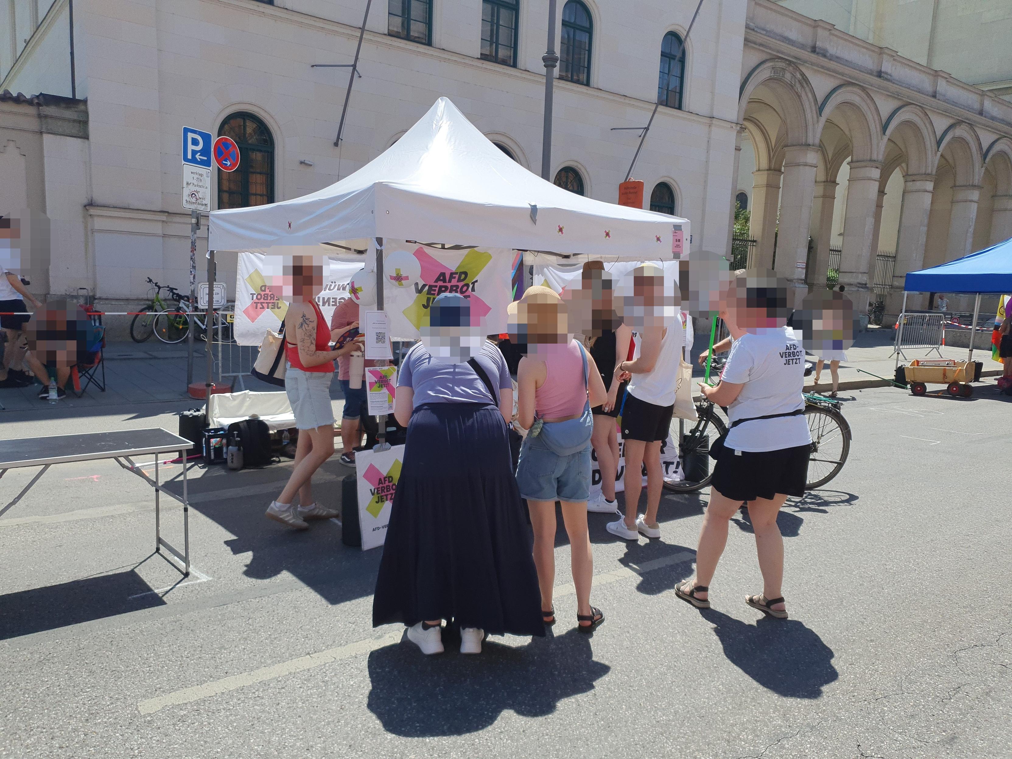 Foto vom AfD-Verbot Jetzt Infostand auf der Pride-Meile beim CSD. Vor einem weißen Pavillon, der mit dem markanten Logo von AfD-Verbot Jetzt! markiert ist, stehen einige Interessierte.