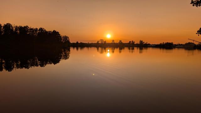 Sonne steht orangerot über einem See und spiegelt sich im spiegelglatten Wasser. Bäume und Büsche spiegeln sich ebenfalls.