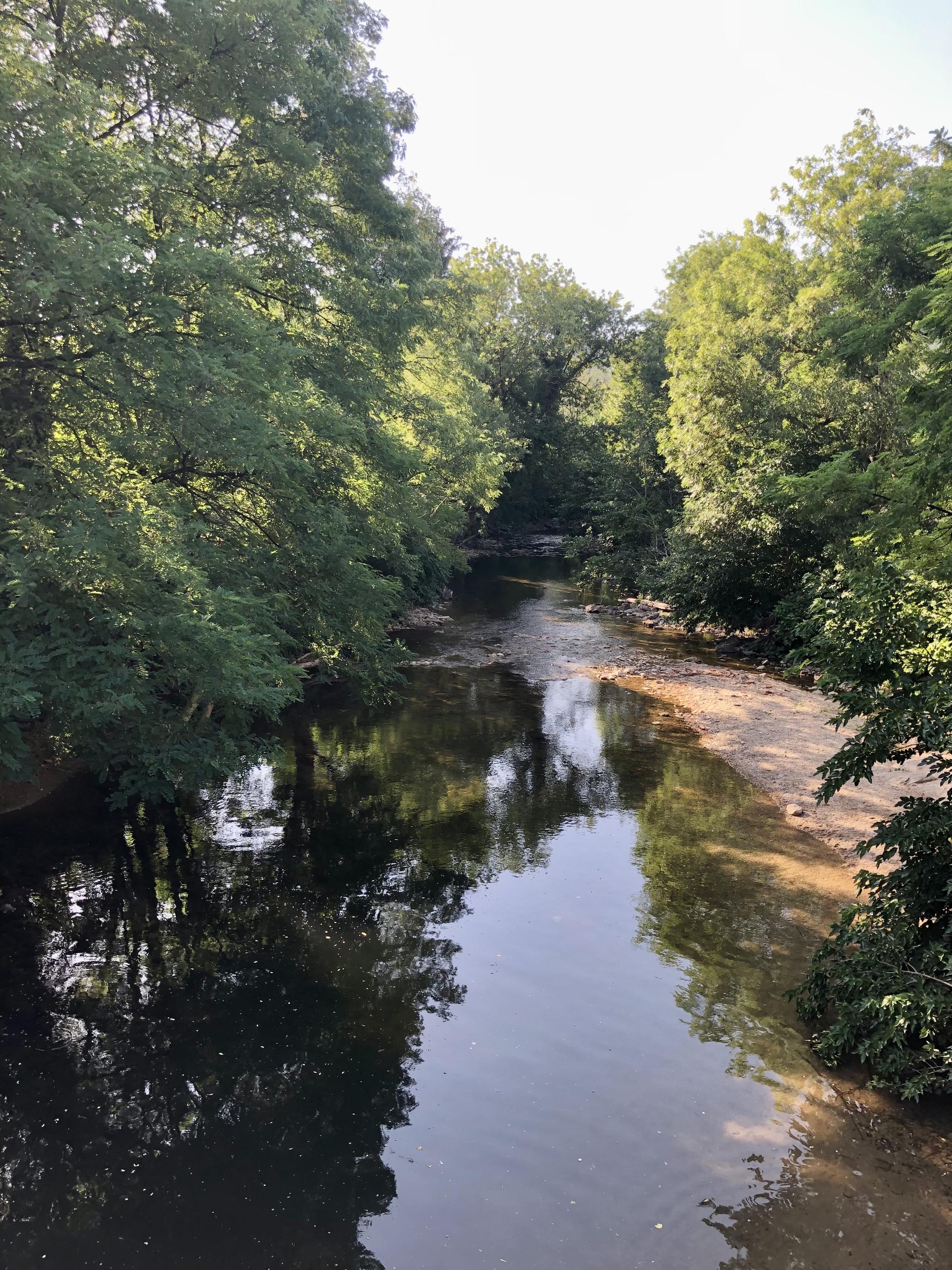 Blick von einer Brücke auf einen kleinen Fluss mit Badestrand. Links und rechts Bäume.