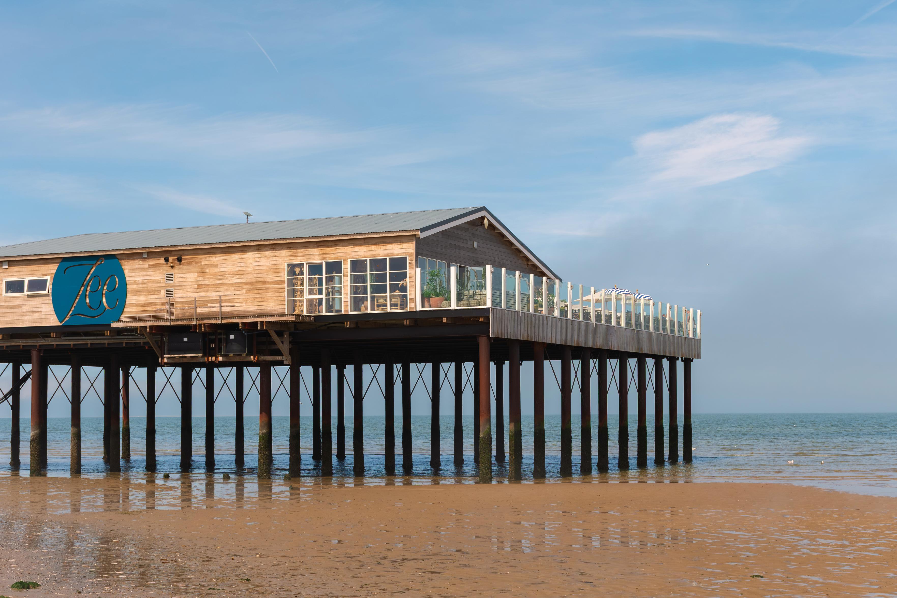 Das Bild zeigt ein Holzgebäude, das auf Stelzen über einem sandigen Strand und dem Meer steht. Der Himmel ist blau mit einigen weißen Wolken. Das Gebäude hat eine Holzfassade und ein Metalldach. An der Vorderseite des Gebäudes befindet sich ein großes blaues Logo mit der Aufschrift "Zee". Es gibt große Fenster, die auf das Meer blicken, und eine Terrasse mit Geländer. Die Stelzen, auf denen das Gebäude steht, sind aus Holz und ragen aus dem Wasser. Der Strand ist nass und reflektiert die Stelzen und das Gebäude. Das Meer ist ruhig und blau. Das Bild vermittelt ein Gefühl von Ruhe und Entspannung. Es könnte ein Restaurant oder ein Café am Meer sein.