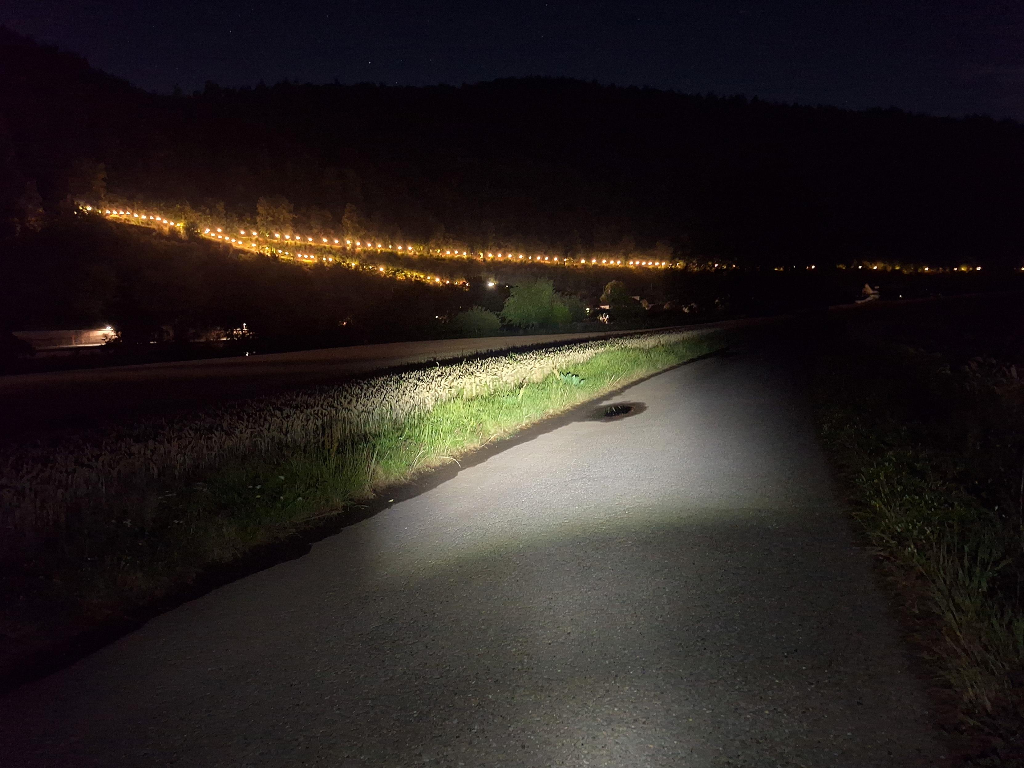 Auf einem dunklen Asphaltweg ist der Schein einer Fahrradleuchte zu sehen. Gegenüber liegen Hügel, teilweise durch Straßenlaternen beleuchtet.