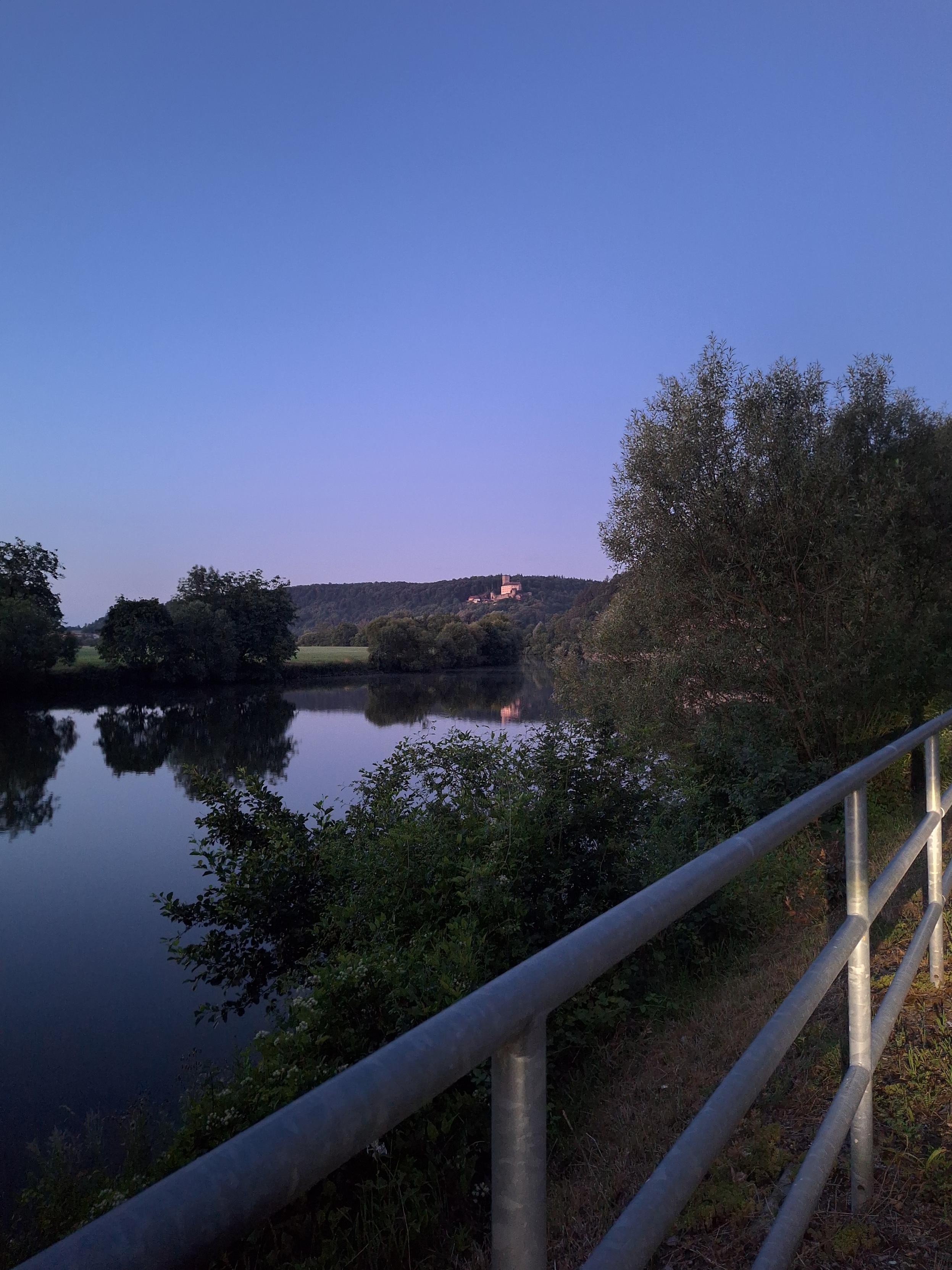 Ein spiegelglatter Fluss in der Morgendämmerung. Ein Geländer im Vordergrund. Eine beleuchtete Burg im Hintergrund.