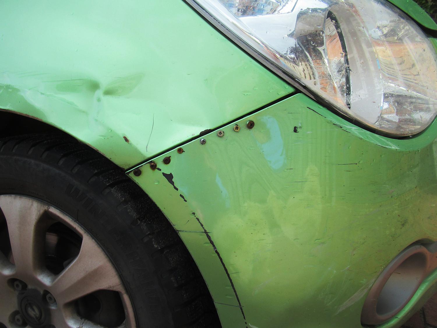 Delle und Kratzern im Bereich vom Kotflügel eines grünen Opel. Die beschädigte Stoßstange wurde provisorisch mit verschiedenen Schrauben befestigt, die bereits Rostspuren zeigen.
