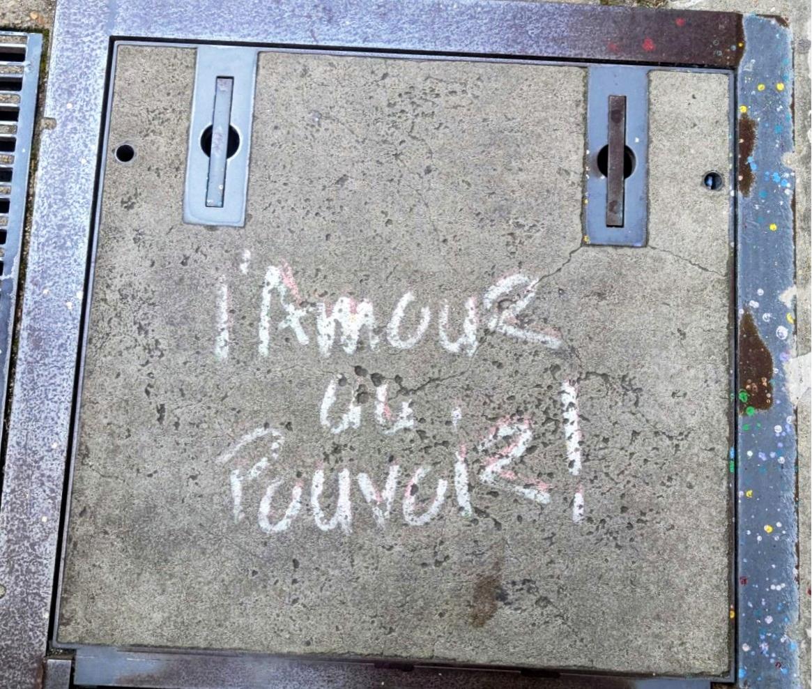 Ein quadratischer Kanaldeckel mit metallenen Einfassungen auf der Straße. In der Mitte des Deckels steht handschriftlich mit weißer Farbe: l'Amour au Pouvoir!