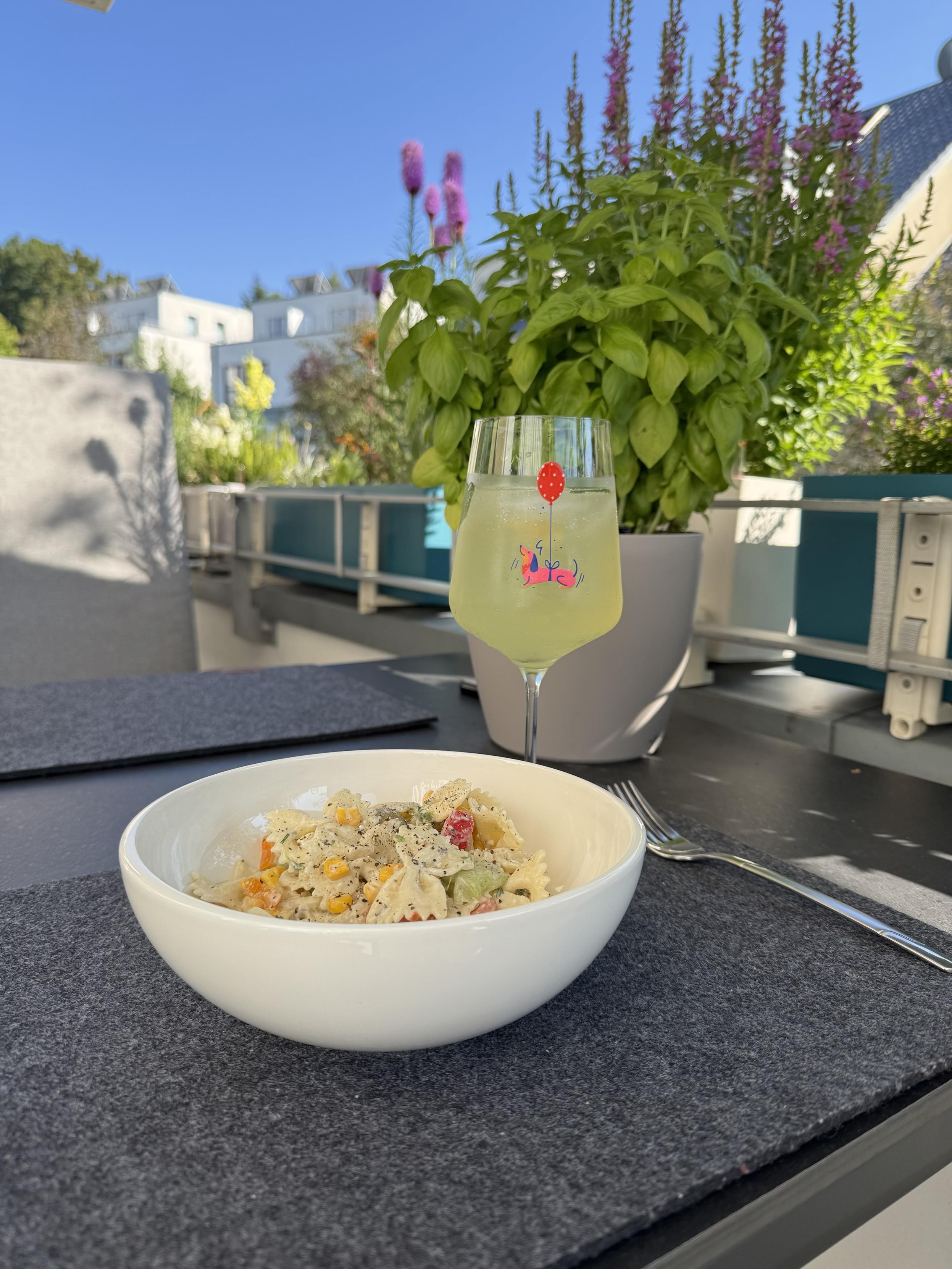 A bowl of colorful pasta salad is placed on a grey placemat, accompanied by a drink served in a glass with a playful design. The background features vibrant flowers and greenery under a clear blue sky, suggesting an outdoor setting.